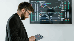 Homme en costume consultant une tablette avec graphique financier devant un écran de trading